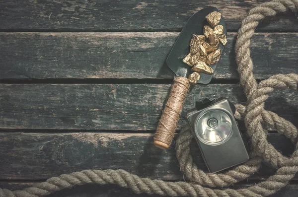 Gold nuggets on the spade, rope and flashlight on the aged wooden table background with copy space. Treasure hunter or goldminer concept.