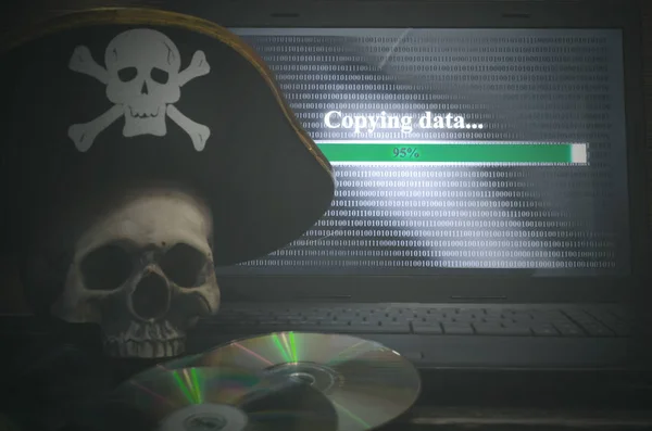 Illegal data copying concept. Cybercrime. Computer piracy background. Pirate hat, human skull, laptop and compact disc on a table.