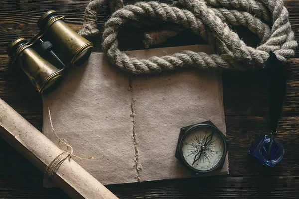 Blank traveler notebook, compass, binoculars, mooring rope and inkpot with a quill pen on a wooden table background mockup. Adventurer notes.