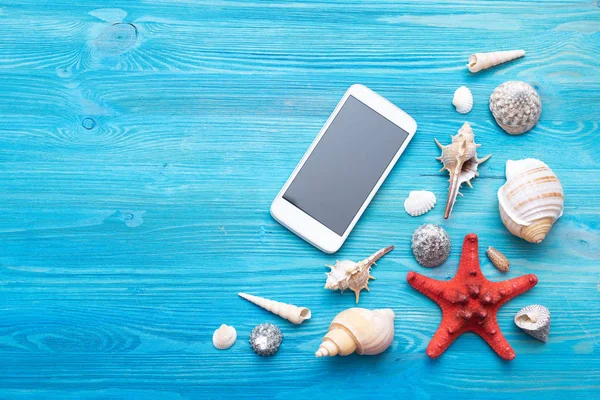 Summer sea vacation concept background with copy space. Blank screen mobile phone and various seashells and a red starfish on a blue wooden floor background.