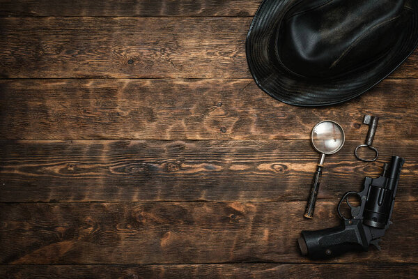 Detective or spy agent wooden table flat lay background with copy space. Handgun, black leather hat, magnifying glass and a rusty key on the work table.