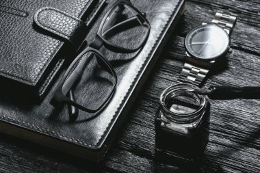 İş geçmişi. Siyah deri kitap, tükenmez kalem, cüzdan, gözlük ve siyah ahşap masa üzerinde bir el saati..