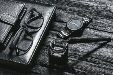 İş geçmişi. Siyah deri kitap, tükenmez kalem, cüzdan, gözlük ve siyah ahşap masa üzerinde bir el saati..