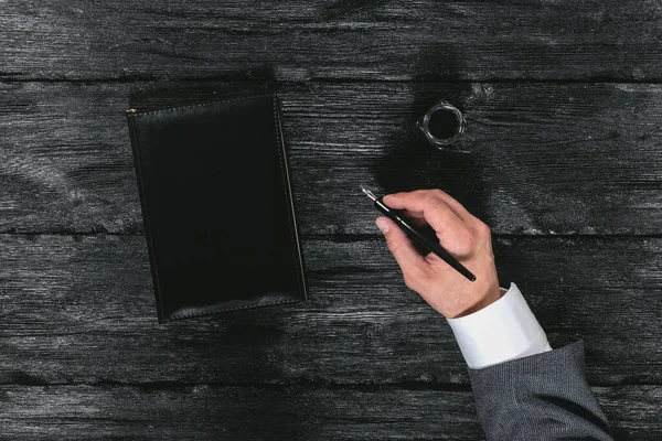 Businessman hand with a quill pen and black cover book on the black wooden table background.