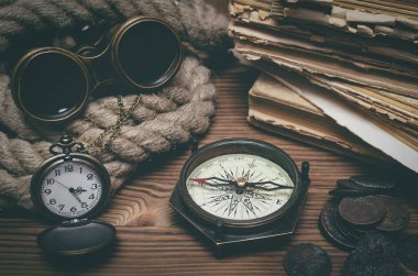 Seyahat ya da macera geçmişi. Macera masası. Dürbünler, eski kitaplar, demirleme halatı, pusula, antik paralar ve masanın üzerinde bir cep saati..