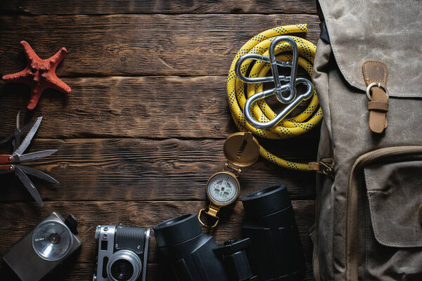 Travel or adventure flat lay background with copy space. A backpack, photo camera, flashlight, multitool knife, rope, carbines, binoculars, compass and a starfish on a brown wooden table.