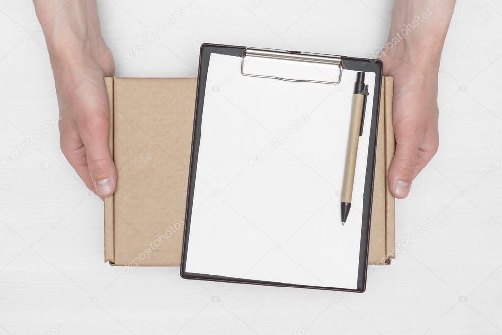 A parcel cardboard box in a delivery boy hands and a blank invoice document mock up on a white wooden table background. Delivery service concept.