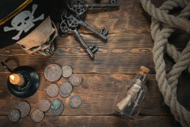 Pirate letter parchment in a bottle, human skull, old coins, moorings, keys and burning candle on a brown wooden table background. clipart