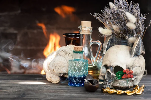 Colorful magic potion bottles and human skull on wooden table on burning fire in fireplace background. Witch doctor table.