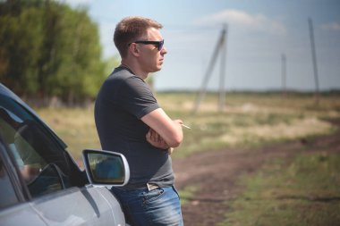 Şoför bir arabanın yanında duruyor ve sigara içiyor..