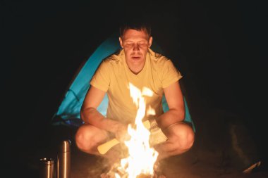 Baltalı yorgun turist yanan bir kamp ateşinin yanında oturuyor..