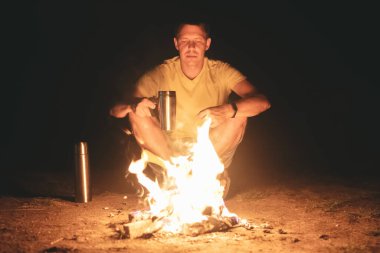 Turist kamp ateşinin yanında oturuyor ve fincandan çay içiyor..