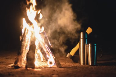 Kamp ateşi, çelik bardak, vakum tüpü ve gece arka planda balta..