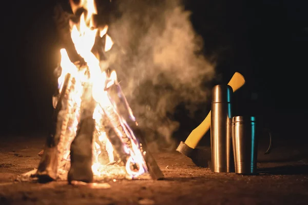 Campfire, steel cup, vacuum flask and axe at night background.