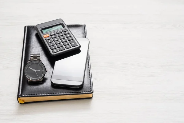Black leather ledger book, calculator, mobile phone and wrist watch on white flat lay background with copy space.