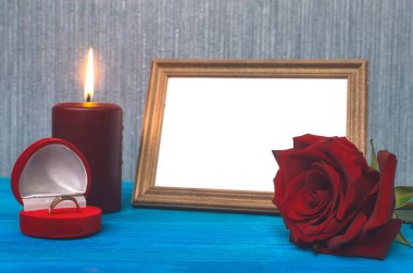 Wedding ring in a gift present box and empty photo frame of a loved one copy space and red rose flower on wooden table background. Marriage offer template. The proposal concept. clipart