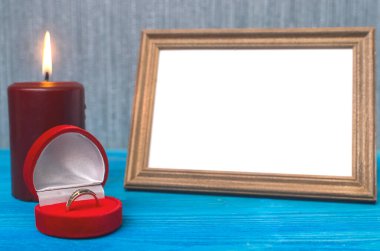 Wedding ring in a gift present box and empty photo frame of a loved one copy space and burning candle on wooden table background. Marriage offer template. The proposal concept. clipart
