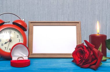 Wedding ring in a gift present box and empty photo frame of a loved one copy space and red rose flower on wooden table background. Marriage offer template. The proposal concept. clipart