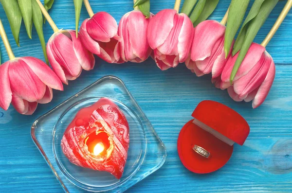 Tulip flowers and wedding ring in a gift present box and burning heart shape candle on blue wooden table board background. Marriage offer. Saint Valentines day romantic backdrop. The proposal.