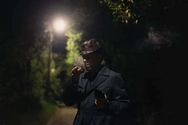 Detective man in the hat and coat with a gun is smoking a smoking pipe in the dark park.