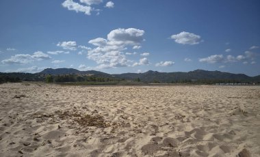 Sardunya 'da bir kumsalın görüntüsü. Gün ışığında bulutlarla kaplı gökyüzünün altında görkemli bir dağ manzarası..