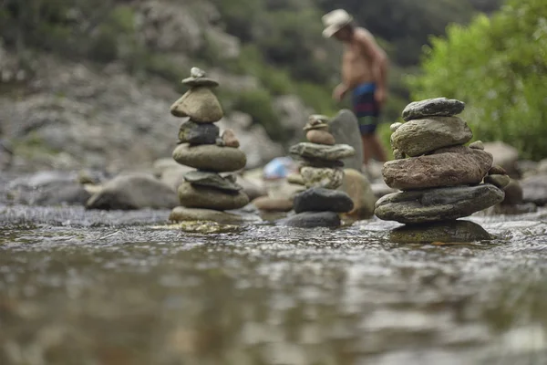 Stacks of zen rocks in a mountain sink with a disfigured man in the background that crosses the stream itself.