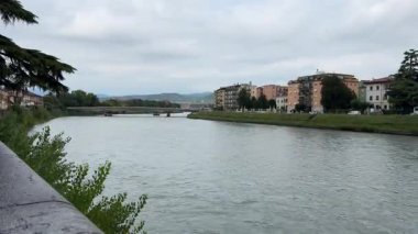 Verona, İtalya 10 Eylül 2025: sakin adige nehri bulutlu bir gökyüzü altında akar, nehir kıyısında binalar ve köprüler görülebilir, verona, İtalya 'da toprak toprak toprak toprak toprak seti yakınlarında az sayıda ağaç ve çalı yetişmektedir.