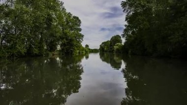 Sakin nehir suları yemyeşil ağaçları ve bulutlu gökyüzünü yansıtıyor. Huzurlu yansımaları ve huzurlu manzara elementleriyle el değmemiş doğal güzellikleri ortaya çıkarıyor.