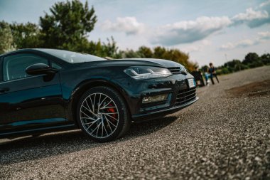 Milan, İtalya 20 Eylül 2025: Siyah Volkswagen Golf r hatchback aracı bulutlu bir gökyüzünün altındaki çakıllı bir yolda duruyor, tasarımı ve kırmızı fren kalibreli spor tekerleği sergiliyor.