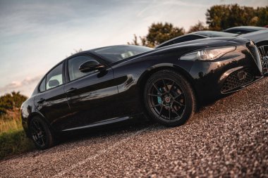 Milan, İtalya 20 Eylül 2025: Alfa Romeo Giulia, günbatımında çakıllı bir yüzeye park edilmiş lüks bir spor sedan, parlak siyah vücuduna sıcak bir ışık yansıtıyor.