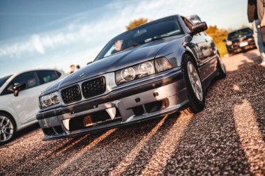 Milan, İtalya 20 Eylül 2025: Gri bmw e36 'lı vampir dişleri açık bir araba yarışında tamponda, sıcak güneş ışığı çakıl yüzeyinde uzun gölgeler bırakıyor.