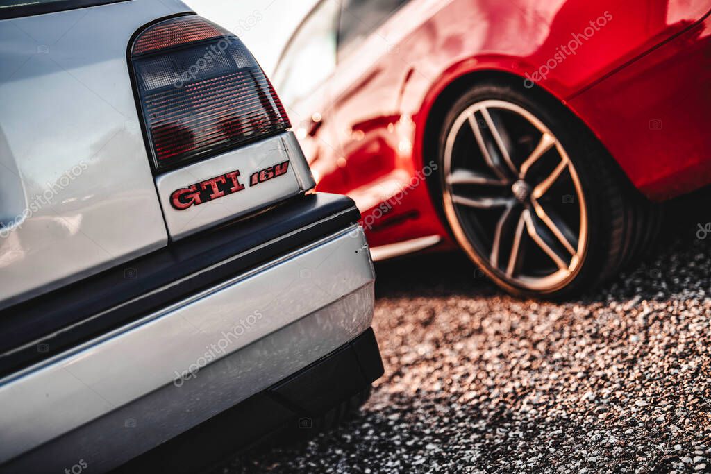Milan, italy 20 september 2025: volkswagen golf gti 16v emblem on a classic silver car bumper, contrasting with a modern red car wheel in the background, parked on gravel