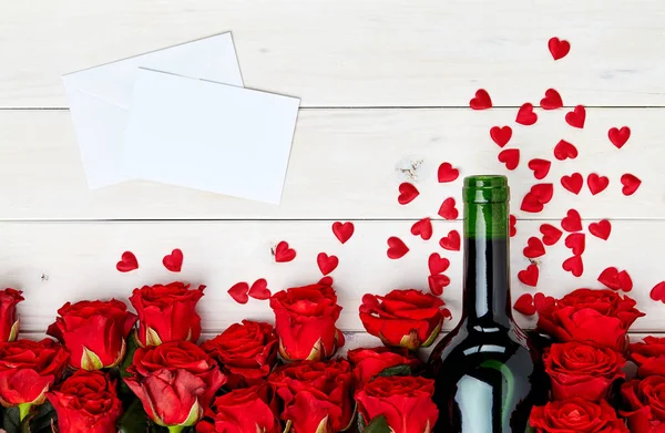Buds of red roses and bottle of wine on white wooden background with confetti and message