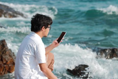 Deniz kıyısı kayaya oturan mobil akıllı telefon olan Genç Asyalı adam. Yaz yaşam tarzı kavramı.