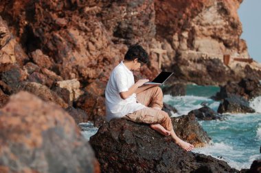 Genç Asyalı adam gezgin deniz kıyısı kayada dizüstü bilgisayar kullanarak. Tatil yaz konsepti.