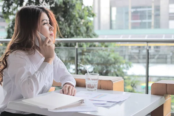 コピー領域の背景が付いているオフィスの机の上のパートナーシップに対してモバイル スマート フォンで話している魅力的な若いアジア ビジネス女性の側面図 — ストック写真