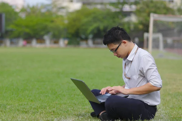 Rahat genç Asyalı iş adamı yeşil çimlerde dizüstü bilgisayarla çalışıyor.