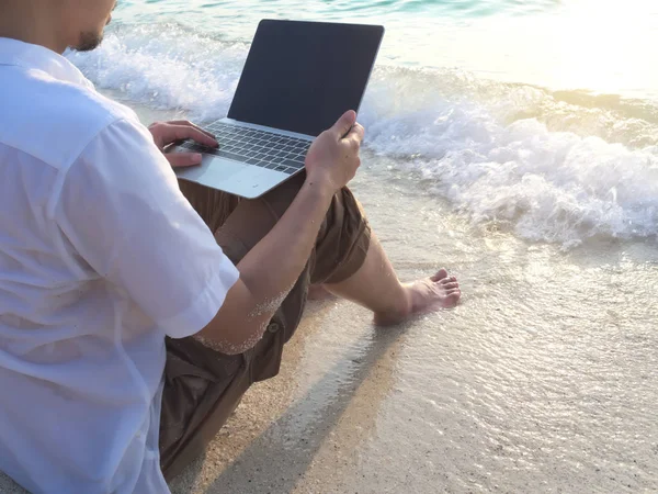 Relaxed young Asian man with laptop sitting on sand of tropical beach. Summer vacations concept.