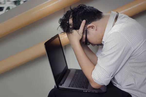 Troubled stressed Asian business man with laptop covering face with hands on staircase of office.