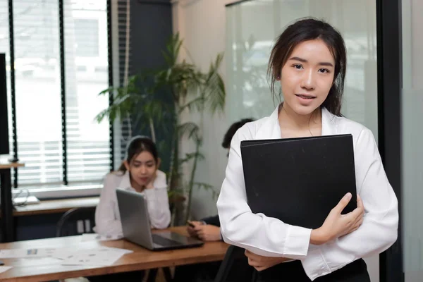 Концепция лидерства среди деловых женщин. Веселая молодая азиатская бизнесвумен с кольцом папки стоя против своего коллеги в офисе фоне . — стоковое фото