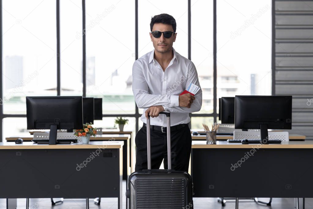 Portrait of cheerful business man holding passport and luggage in workplace of office. Summer vacations concept.