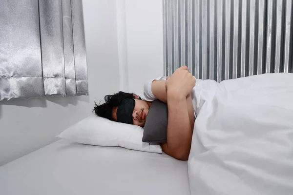 Young Asian man with blindfold eye mask sleeping on the white bed in bedroom.