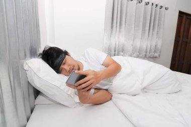 Genç Asyalı adam yatak odasındaki beyaz yatakta cep telefonu kullanıyor. Teknoloji, internet, iletişim ve insan kavramı 