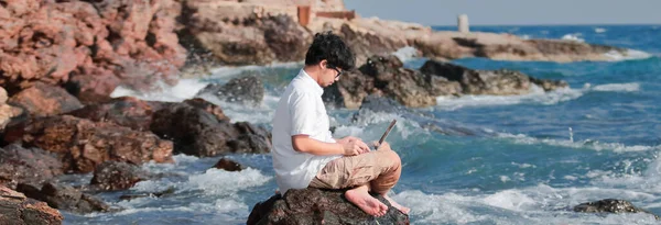 Панорамный Вид Расслабленного Молодого Азиата Ноутбуком Сидящего Скале Берегу Моря — стоковое фото