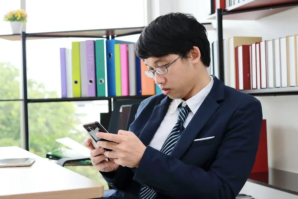 Young Asian man in suit looking mobile smart phone in hands in office.