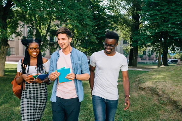 Grupo de tres adolescentes hipster multiétnicos estudiantes amigos riendo y hablando mientras pasan tiempo juntos al aire libre en el parque o campus universitario en la mañana soleada . — Foto de Stock