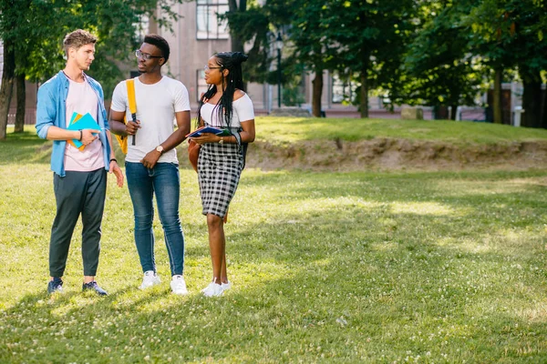 Longitud completa de estudiantes multiétnicos universitarios felices hablando juntos durante el descanso en el soleado día de verano en el campus al aire libre. Copiar espacio. Diferente cultura concepto de amistad — Foto de Stock