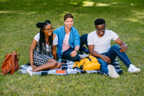 Groupe de trois jeunes étudiants heureux et souriants assis ensemble sur le campus universitaire de l'école secondaire Green Lawn. Concept d'amitié culturelle différente — Photo