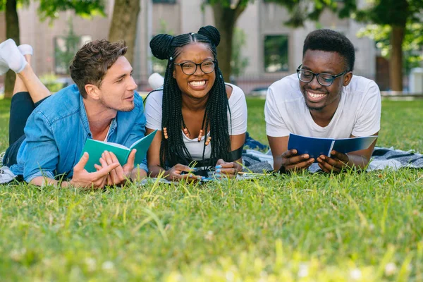 Grupo multiétnico de estudiantes amigos tumbados en el césped hablando riendo leyendo libros mientras hacen la tarea en el parque en el prado soleado. Concepto Educación y Amistad. Mujer negra riendo de broma divertida . — Foto de Stock