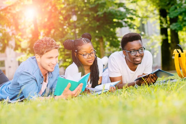 Vooraanzicht van drie gelukkige multi-etnische studenten liggend, praten, samen grapje samen op de Green Lawn High School University campus. Onderwijs en vriendschap concept. Zonneschittering effect — Stockfoto